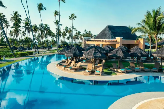 Piscina com ilha de espreguiçadeiras e guarda-sóis de palha diante de um bar com telhado de piaçava